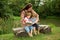 Charming Mother Reading a Book to Her Adorable Little Twin Sons While Sitting Outside Near Beautiful Lake