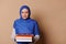 Charming Muslim woman in blue hijab, holding pile of books and smiling looking at camera, on isolated beige background