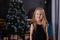 charming preschool blonde girl in a black dress standing near a christmas tree with blue decorations in dark room