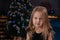 charming preschool blonde girl in a black dress standing near a christmas tree with blue decorations in dark room