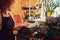 Charming redhead female florist wearing uniform works with cash register in flower shop.