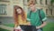 Charming redhead woman joining concentrated Caucasian man sitting on bench on campus and using laptop. Portrait of two