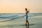 Charming slender blonde in stylish dress stands in waves on the sea. Young woman walks barefoot along the surf and enjoys the rest