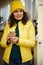 Charming stunning positive multi-ethnic brunette woman, in yellow hat and jacket, with takeaway hot coffee in paper cup