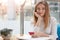 Charming woman with beautiful smile holds mobile phone in hand, keeps another hand on chin, has rest in coffee shop. Caucasian