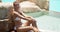 Charming woman in white swimwear leaning on stone nearby pool and smiling at camera