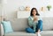 Charming young Indian woman sitting on sofa with smartphone, checking social media, chatting online at home