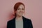 Charming young red-haired female smiling pleasantly at camera, isolated on rosy background