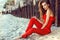 Charming young woman with long hair in coral red one shoulder jumpsuit sitting on the beach at the old rusty piles