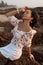 Charming young woman in white skirt and blouse is sitting on ocean shore on rocks, raising her face to setting sun.