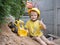 Cheerful 5-year-old boy in yellow T-shirt and in construction helmet plays with a toy bulldozer at construction site