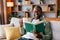 Cheerful adult african american man in glasses and casual reading book and enjoy favorite drink on sofa