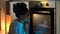 Cheerful African American woman sits near oven in kitchen waiting for delicious cake to be prepared