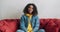 Cheerful african american woman in yellow hoodie and jeans jacket sitting on red sofa at home looking at camera and