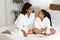 Cheerful African Girl And Her Mother In Bathrobes Reading Book On Bed