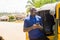 cheerful african man standing next to his tuk tuk taxi smiling and using his smart phone