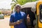 cheerful african man standing next to his tuk tuk taxi smiling and using his smart phone giving a thumbs up