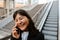 Cheerful asian girl talking on smartphone while standing on escalator outdoors