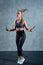 Cheerful athletic girl in sportswear woman training with a skipping rope in a gym on a grey wall.