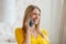 Cheerful attractive millennial blond european female in yellow clothes speaks by phone in room interior