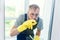 Cheerful bearded man in funny eyeglasses cleans the window with detergents and remove dirt with his finger in gloves.