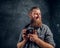 A cheerful bearded photographer wearing a gray holding a photo camera and looking at a camera.