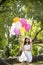 Cheerful beauty woman holding balloons relax sitting under big tree in green park with happiness. Woman Hands holding vibrant air