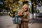 Cheerful blond woman in coat holds basket with flowers inside.