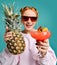 Cheerful blonde woman in modern red sunglasses handed us demonstrates a strawberry margarita cocktail and a big pineapple