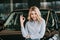 Cheerful blonde woman showing ok sign near black automobile in car showroom.