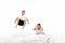 Cheerful boy levitating over shocked brother sitting on bedding and holding hands on head