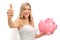 Cheerful bride making a thumb up sign and holding a piggybank