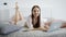 Cheerful brown haired woman is lying on a bed in room in day with notebook in front of her and looking to a camera