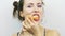 Cheerful brunette girl eating mandarin at camera against white background. Smiling young woman with pink lips eats orange at camer