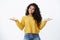 Cheerful carefree alluring african-american woman with curly dark hairstyle in yellow sweater, spread hands sideways