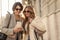 Cheerful caucasian adult women look into phone screen while sharing information outdoors.