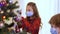 Cheerful Caucasian girl in coronavirus face mask decorating Christmas tree with brother in living room at home. Portrait