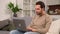 Cheerful caucasian man wearing casual wear sitting on sofa and using laptop at modern home office