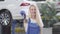 Cheerful Caucasian woman standing at the background of car and showing car keys to camera. Blond female auto mechanic