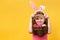 A cheerful child in the Easter ears of a bunny peeks out from under the handle of a basket. Little girls on a yellow background