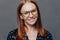 Cheerful close up shot of pretty woman with brown hair, toothy smile, wears spectacles, dressed in formal blouse, poses over grey