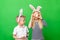 Cheerful crazy boy and girl in rabbit bunny ears on head on green studio background. Easter painting eggs on eyes
