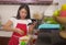 Cheerful domestic cooking - young happy and beautiful Asian Korean home cook woman in red apron reading healthy recipe in mobile