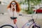 Cheerful female urban biker wears sunglasses and dress, has luxurious crisp foxy hair, keeps elbows on saddle of bicycle, spends