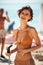 Cheerful girl in bikini using hose pipe on beach. Portrait of smiling lady in beige swimsuit rinsing beach sand off her