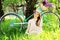Cheerful girl.Horizontal shot of beautiful young woman in dress and hat adjusting hat and smiling while sitting on grass with bike