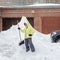 Cheerful girl with the shovel for snow removal stands near a huge snowdrift near the garage