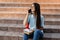 A cheerful girl is talking on a mobile phone with her friend, sitting with a notebook on the stairs in the campus