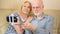 Cheerful good-looking senior couple sitting on sofa at home. Making selfie with smartphone