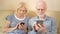 Cheerful good-looking senior couple sitting on sofa at home. Using smartphone, browsing, reading